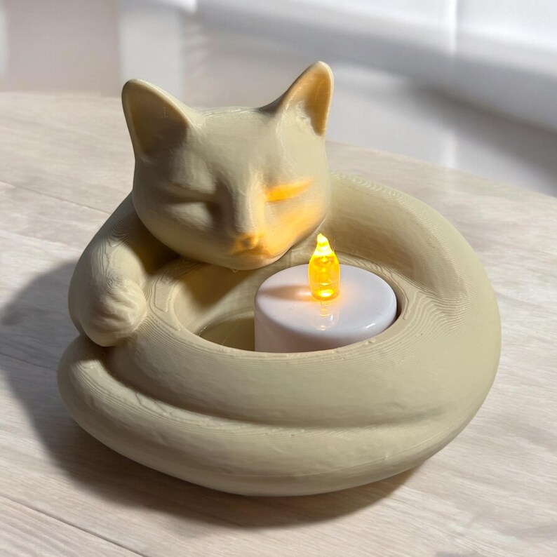 May include: A beige cat-shaped candle holder with a sleeping cat design. The cat's body forms a circular base, holding a white candle with a yellow LED flame. The candle holder is made of ceramic.