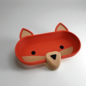May include: A whimsical orange fox-shaped tray with tan accents. The tray features black circular eyes and a triangular nose. The ears are tan with orange outlines. The tray is designed with a rounded rectangular shape.
