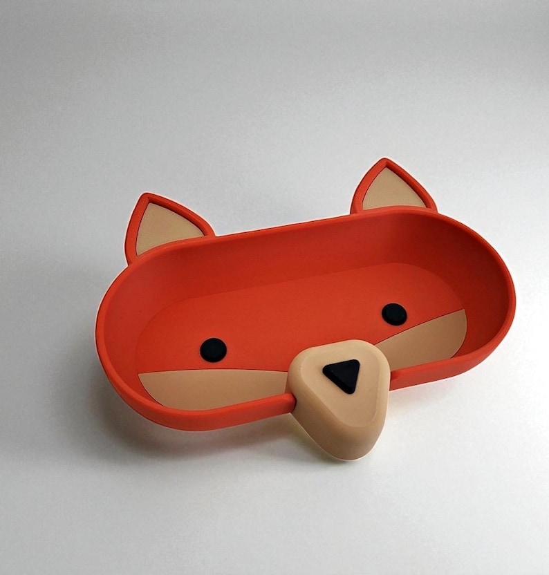 May include: A whimsical orange fox-shaped tray with tan accents. The tray features black circular eyes and a triangular nose. The ears are tan with orange outlines. The tray is designed with a rounded rectangular shape.