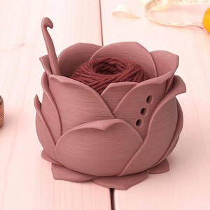 May include: A dusty rose-colored yarn holder shaped like a rose, with a ball of brown yarn inside. A crochet hook is placed in the holder. The holder has three small holes on the side. The holder is sitting on a light pink surface.