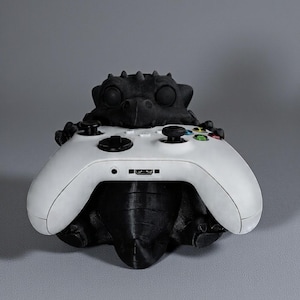 May include: A black dragon-shaped controller stand holding a white Xbox controller. The dragon has a textured, scaly design and is 3D-printed. The controller has black joysticks and coloured buttons.