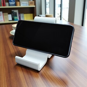 Op de afbeelding: Een witte, geribbelde telefoonstandaard die een zwarte smartphone ondersteunt. De standaard is gemaakt van een stevig materiaal en heeft een gebogen ontwerp. De standaard staat op een houten tafel. De standaard is ongeveer 10 cm lang.