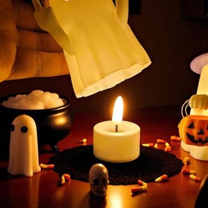 May include: A Halloween-themed display featuring a lit candle, a small skull, and a miniature ghost figure. A cauldron with white foam and candy corn are also present. A large ghost-shaped lamp is held above the scene.