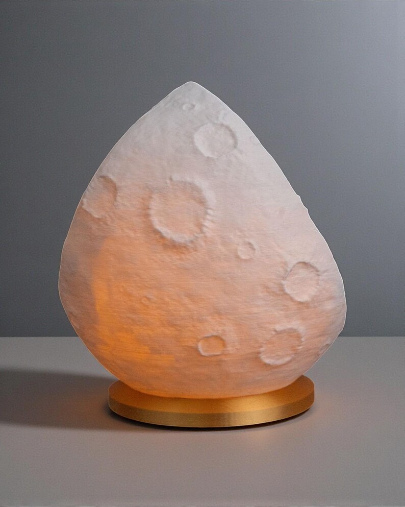 May include: A white, teardrop-shaped lamp with a textured surface and crater-like indentations. The lamp sits on a round, gold-colored base. The interior glows with a warm, inviting light, creating a soft ambiance. A decorative lighting fixture.