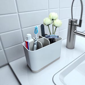 以下が含まれることがあります： 歯磨き粉のチューブ、歯ブラシ、爪やすり、カミソリ、白い花3本が入った白とベージュのバスルームオーガナイザー。 オーガナイザーは、洗面台と蛇口の近くの白いカウンタートップに置かれています。