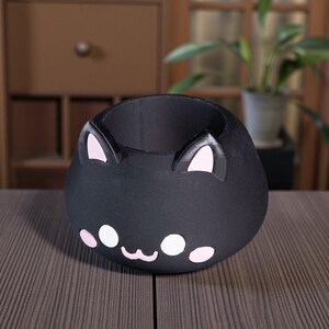 K&ouml;nnte beinhalten: Ein schwarzer Katzen-Blumentopf mit rosa Akzenten. Der Topf hat einen runden K&ouml;rper mit einem l&auml;chelnden Gesicht, rosa Ohren und wei&szlig;en und rosa Gesichtsdetails. Er steht auf einer braunen Oberfl&auml;che.