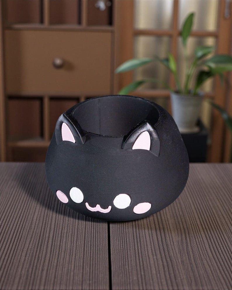 K&ouml;nnte beinhalten: Ein schwarzer Katzen-Blumentopf mit rosa Akzenten. Der Topf hat einen runden K&ouml;rper mit einem l&auml;chelnden Gesicht, rosa Ohren und wei&szlig;en und rosa Gesichtsdetails. Er steht auf einer braunen Oberfl&auml;che.