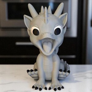 May include: A light gray dragon figurine with large black eyes and an open mouth, revealing a pink tongue. The dragon has a spiky crest, small ears, and black claws. The figurine is sitting on a white surface.