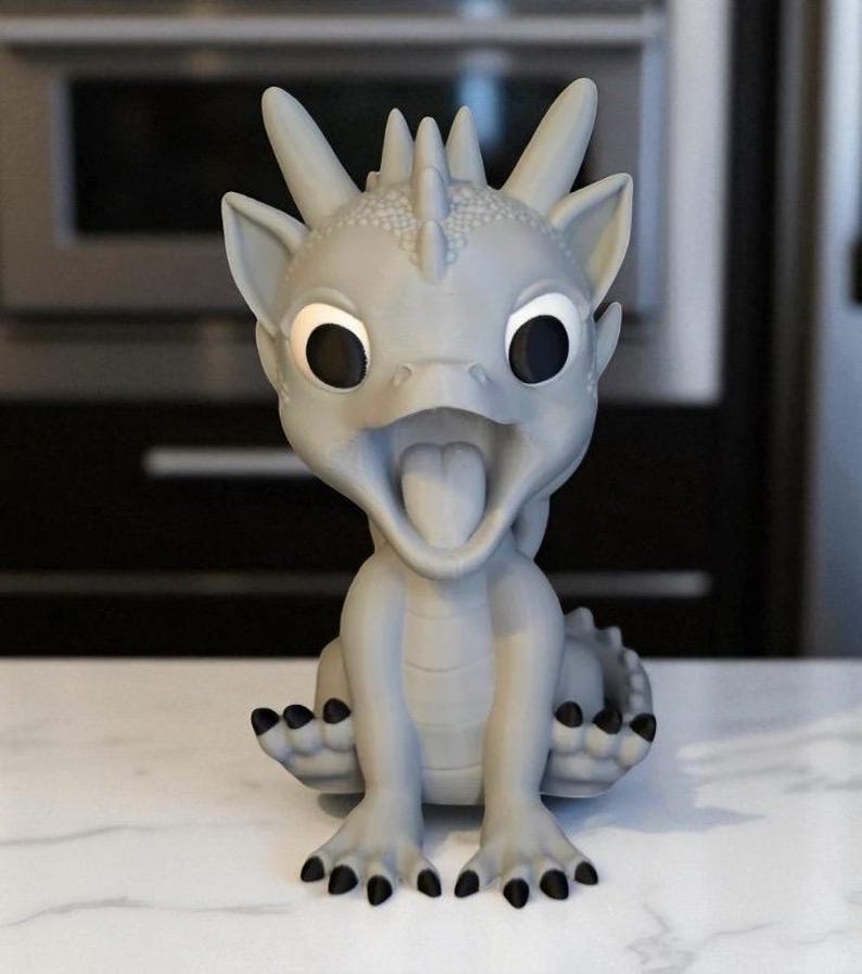 May include: A light gray dragon figurine with large black eyes and an open mouth, revealing a pink tongue. The dragon has a spiky crest, small ears, and black claws. The figurine is sitting on a white surface.