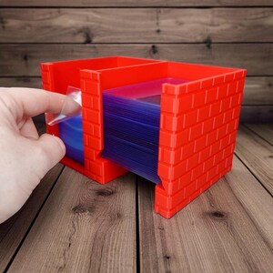 May include: A red card holder with a brick pattern, featuring multiple compartments. A hand is shown placing a card into a clear plastic sleeve. The holder is designed to organise and protect trading cards or similar items.