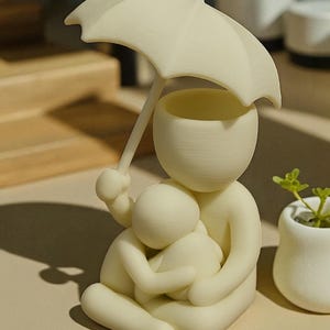 May include: A cream-colored figurine depicts a parent figure holding a child, both sheltered under an umbrella. The umbrella is held by the parent figure. The figurine is sitting down. A small white pot with a green plant is to the right.