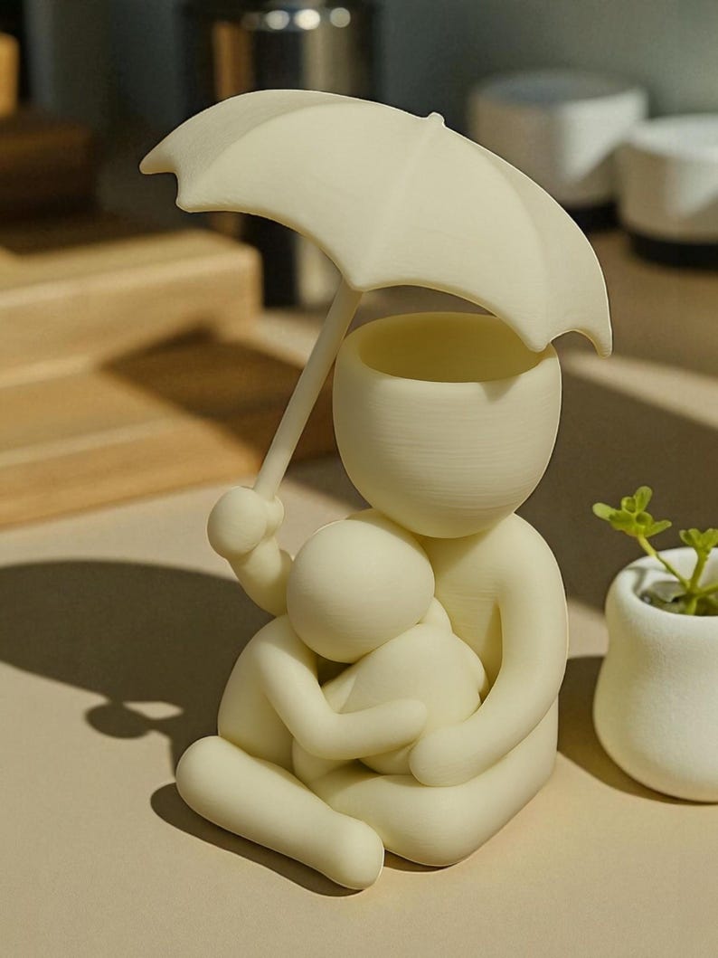May include: A cream-colored figurine depicts a parent figure holding a child, both sheltered under an umbrella. The umbrella is held by the parent figure. The figurine is sitting down. A small white pot with a green plant is to the right.