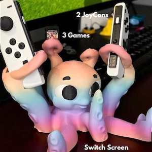 Puede incluir: Un soporte para consola de juegos Switch con forma de pulpo colorido. El soporte sostiene dos Joy-Cons blancos y un cartucho de juego. El texto en la imagen dice "2 JoyCons", "3 Games" y "Switch Screen". El pulpo está pintado en colores pastel.