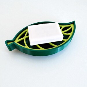 May include: A green leaf-shaped soap dish with a yellow interior, holding a white bar of soap. The dish has a detailed leaf vein design and is made of a solid material. The soap dish is designed to hold a bar of soap.