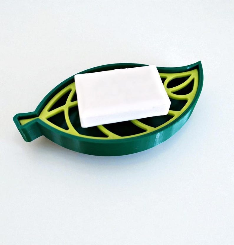 May include: A green leaf-shaped soap dish with a yellow interior, holding a white bar of soap. The dish has a detailed leaf vein design and is made of a solid material. The soap dish is designed to hold a bar of soap.