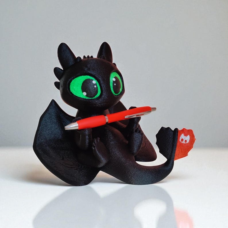 May include: A black dragon figurine with large green eyes, holding a red pen. The dragon has large wings and a tail with a red fin. The figurine is sitting on a white surface.