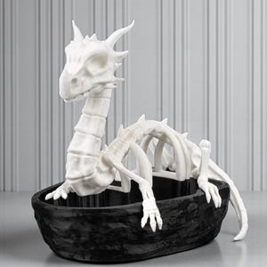 May include: A white dragon skeleton sculpture in a black textured boat-shaped container. The dragon has a detailed skull, spine, and rib cage. The sculpture is a decorative item, possibly for Halloween or fantasy decor.