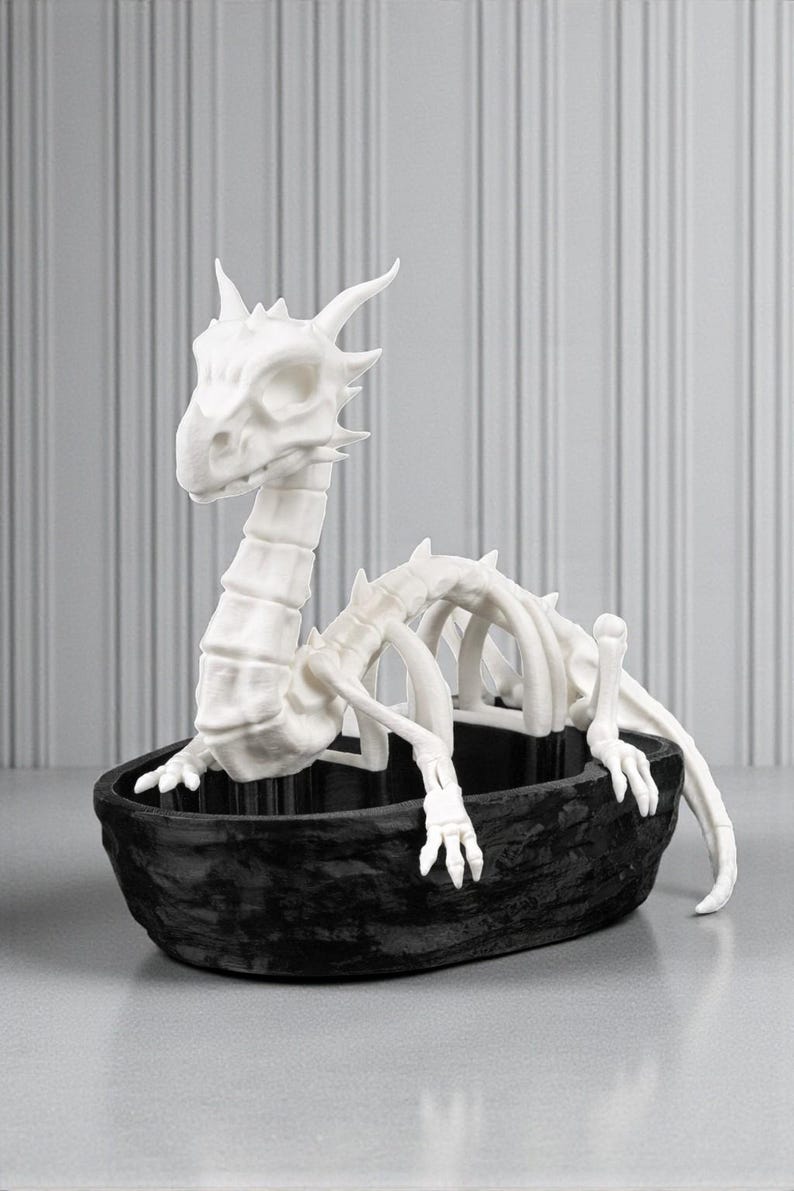 May include: A white dragon skeleton sculpture in a black textured boat-shaped container. The dragon has a detailed skull, spine, and rib cage. The sculpture is a decorative item, possibly for Halloween or fantasy decor.