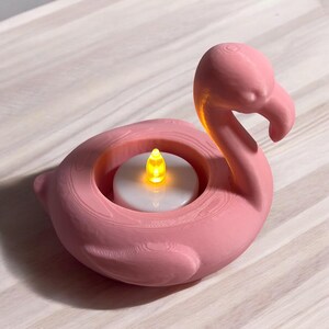 May include: A pink flamingo-shaped candle holder with a glowing yellow LED candle. The holder is made of a textured material and sits on a light-colored wooden surface. The flamingo's head is upright, and its beak is curved.