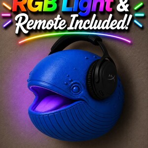 Op de afbeelding: Een blauwe walvisvormige lamp met een gloeiende paarse mond en een zwarte headset. De lamp heeft de tekst "RGB Light & Remote Included!" in kleurrijke letters. De lamp staat op een bruine achtergrond.
