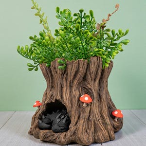 Op de afbeelding: Een decoratieve boomstamplanter met groene bladeren en drie rood-witte paddenstoelaccenten. Een kleine grot herbergt twee zwarte drakenbeeldjes. De planter is gemaakt van een bruin, getextureerd materiaal.