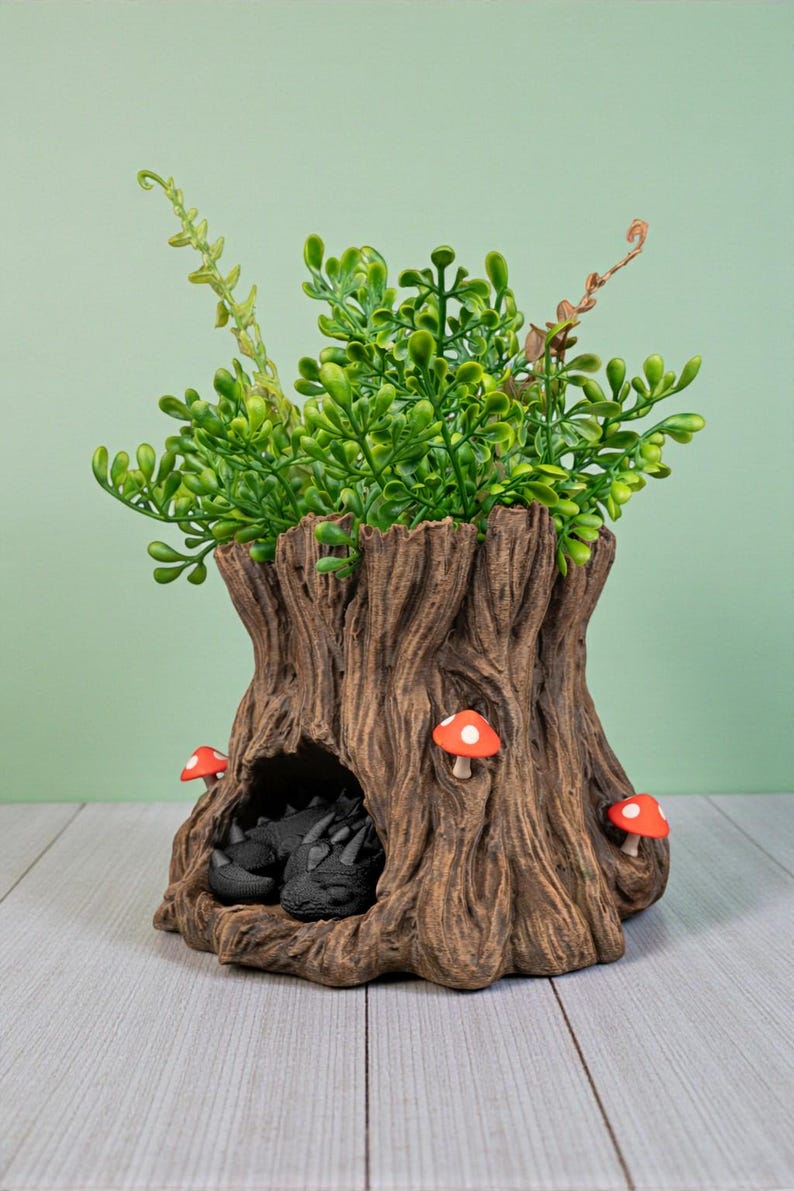 Op de afbeelding: Een decoratieve boomstamplanter met groene bladeren en drie rood-witte paddenstoelaccenten. Een kleine grot herbergt twee zwarte drakenbeeldjes. De planter is gemaakt van een bruin, getextureerd materiaal.
