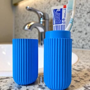 Pode incluir: Dois porta-escovas de dentes azuis e canelados. Um cont&eacute;m um tubo de pasta de dentes e uma escova de dentes. Os suportes cil&iacute;ndricos est&atilde;o sobre uma bancada salpicada. Uma torneira e uma pia prateadas est&atilde;o no fundo.