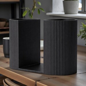 May include: A pair of black metal bookends with a ribbed design. One is rectangular, the other is cylindrical, and they are joined by a flat base. The bookends are on a wooden table, with plants and decor in the background.