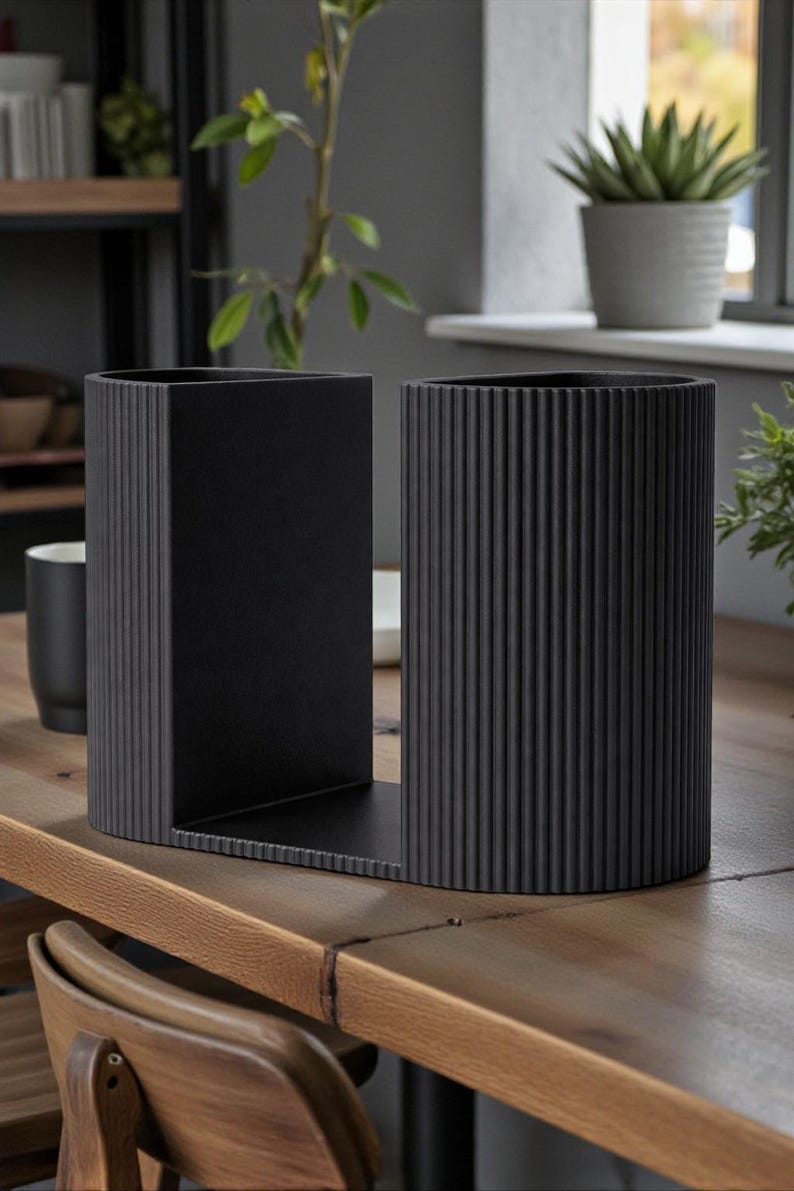 May include: A pair of black metal bookends with a ribbed design. One is rectangular, the other is cylindrical, and they are joined by a flat base. The bookends are on a wooden table, with plants and decor in the background.