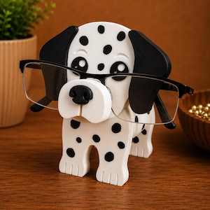 K&ouml;nnte beinhalten: Ein Brillenhalter in Form eines Dalmatinerhundes mit wei&szlig;en K&ouml;rper und schwarzen Flecken und Ohren. Der Halter h&auml;lt eine Brille mit schwarzem Rahmen. Der Hund steht auf einer Holzoberfl&auml;che.
