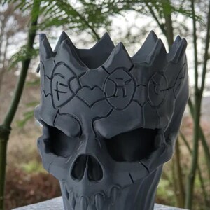 May include: A gray skull-shaped planter with a crown design. The planter has a detailed, textured surface with carved patterns around the crown and face. The skull features eye sockets, a nose cavity, and a row of teeth.