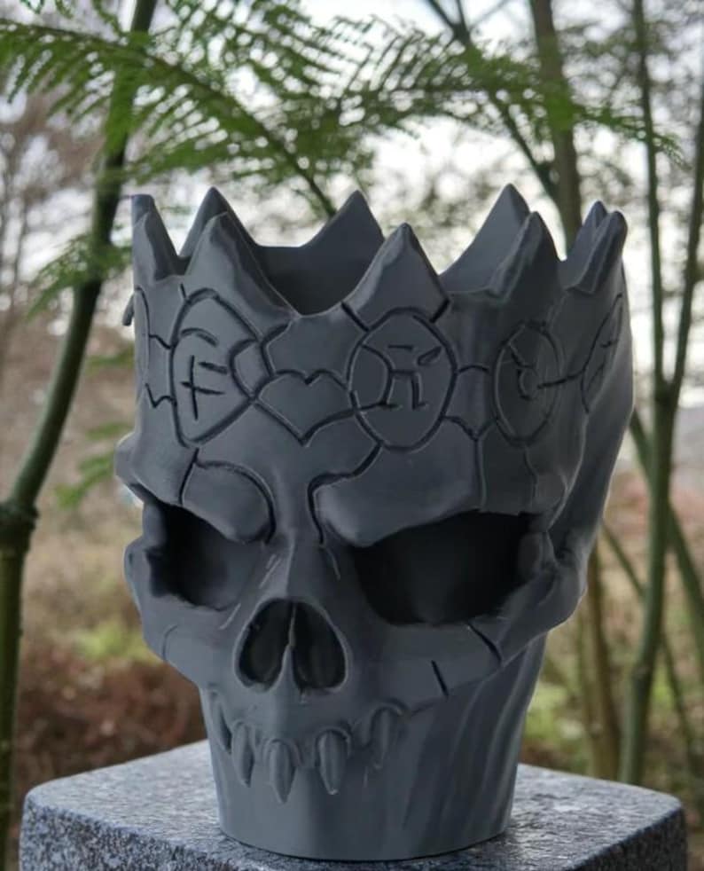May include: A gray skull-shaped planter with a crown design. The planter has a detailed, textured surface with carved patterns around the crown and face. The skull features eye sockets, a nose cavity, and a row of teeth.