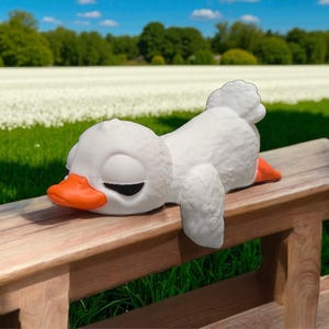 May include: A white, sleeping duck figurine with an orange beak and feet rests on a wooden bench. The duck is lying down with its eyes closed. The background features a field of white flowers and a blue sky.