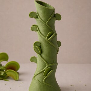 May include: A green, stylized vase with a textured surface, resembling a plant stem. The vase features leaf-like accents and winding vines, creating a botanical theme. The vase is approximately 10 inches tall.