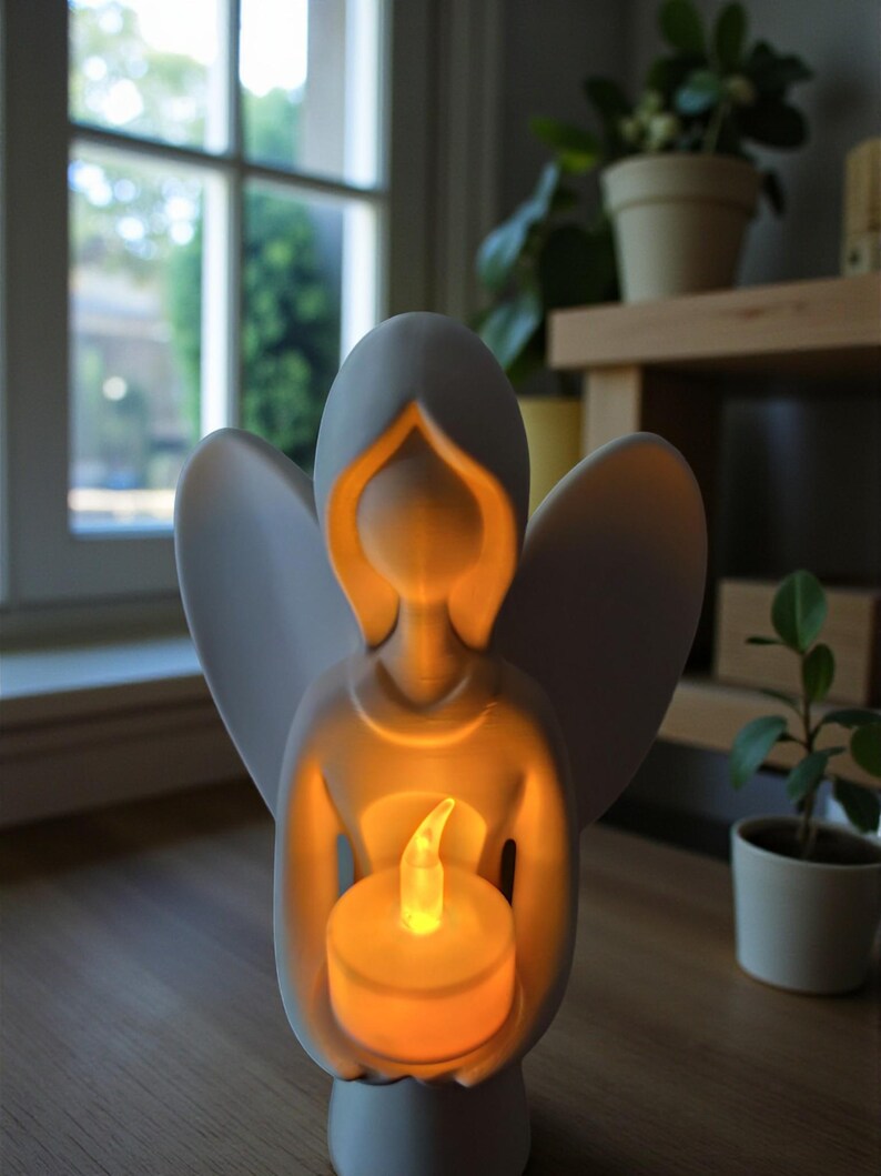 May include: A white angel figurine holding a glowing LED candle. The angel has outstretched wings and a serene expression. The candle emits a warm, inviting light, creating a peaceful ambiance. The figurine is on a wooden surface.