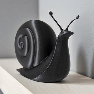 May include: A black snail ornament with a detailed shell and antennae. The sculpture is made of a solid material and is placed on a light wooden shelf. The design is minimalist and modern.