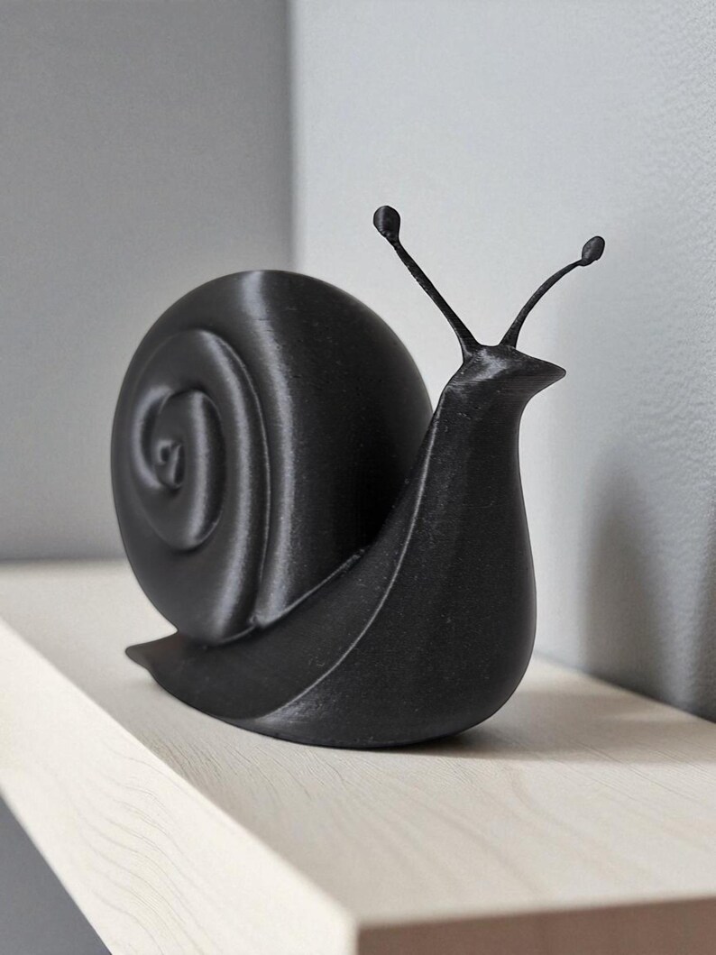 May include: A black snail ornament with a detailed shell and antennae. The sculpture is made of a solid material and is placed on a light wooden shelf. The design is minimalist and modern.