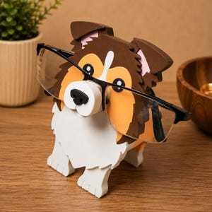 May include: A wooden dog-shaped eyeglasses holder in white, brown, and orange. The dog is wearing black-framed glasses. The holder is on a wooden surface, with a plant in a pot and a wooden bowl in the background.