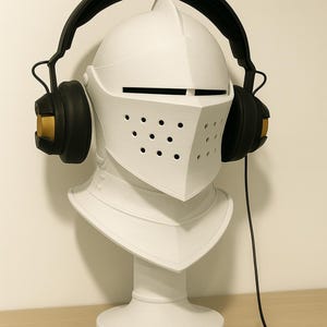 Op de afbeelding: Een witte ridderhelm buste met zwarte koptelefoon. De helm heeft een vizier met kleine gaatjes en een puntige bovenkant. De koptelefoon heeft zwarte oorschelpen met gouden accenten. Een zwarte kabel komt uit de koptelefoon.
