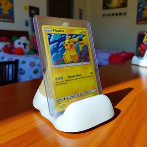 May include: A yellow Pikachu Pok&eacute;mon card in a clear plastic holder, displayed in a white, curved stand. The card features a yellow Pikachu character with the text "Thunder Shock". The stand is on a wooden surface.