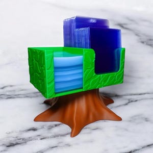 May include: A green and brown card holder shaped like a tree. The green portion has a leaf design and holds blue and clear cards. The base is brown and resembles a tree trunk. The holder is on a white marble surface.