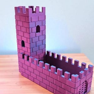 May include: A purple and pink castle-shaped planter with a tall tower and crenelated walls. The planter has a rectangular base and a small arched doorway. The brickwork is detailed, and the color gradient adds depth.