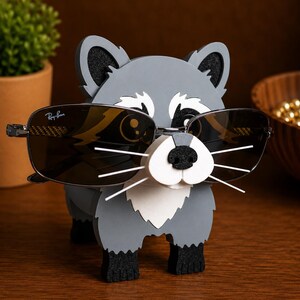 May include: A gray raccoon-shaped eyeglass holder with black accents, holding a pair of Ray-Ban sunglasses. The holder has a white muzzle and whiskers, with black paws and ears. The sunglasses have dark lenses and a silver frame.