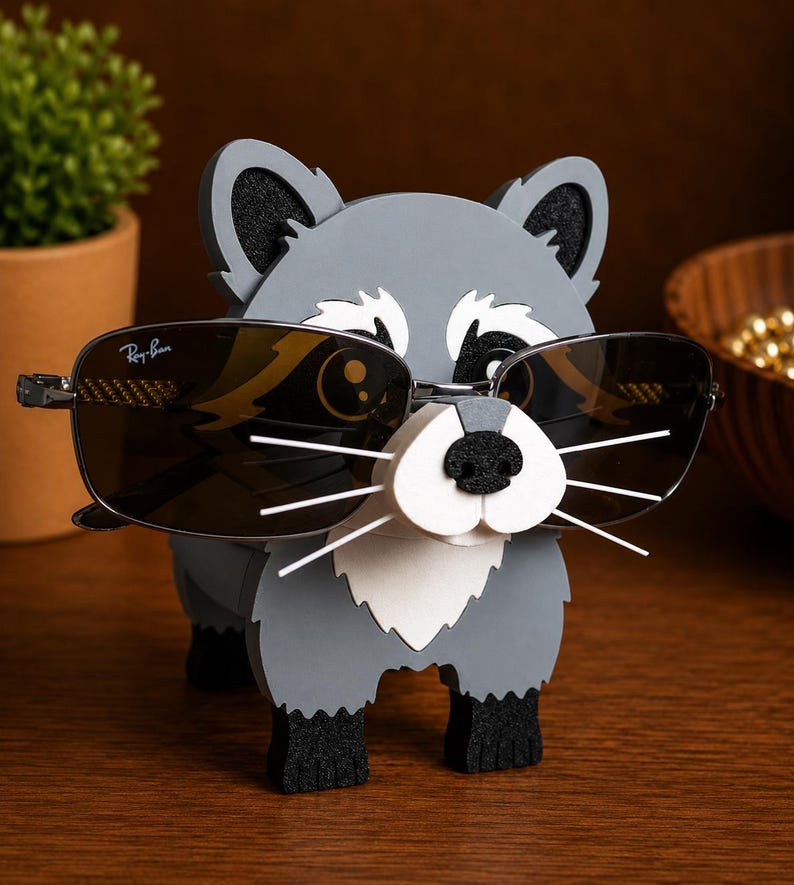 May include: A gray raccoon-shaped eyeglass holder with black accents, holding a pair of Ray-Ban sunglasses. The holder has a white muzzle and whiskers, with black paws and ears. The sunglasses have dark lenses and a silver frame.
