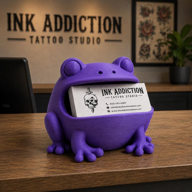 May include: A purple frog-shaped business card holder. The frog has a wide open mouth holding a stack of white business cards. The business cards have the text "INK ADDICTION - TATTOO STUDIO" and a skull graphic. The holder is on a wooden surface.