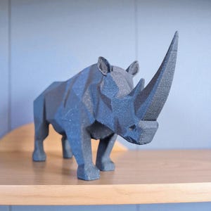 May include: A dark gray, geometric rhino sculpture with a prominent horn. The rhino is positioned on a light brown wooden shelf against a light blue wall. The sculpture's design features sharp angles and a textured surface, giving it a modern aesthetic.
