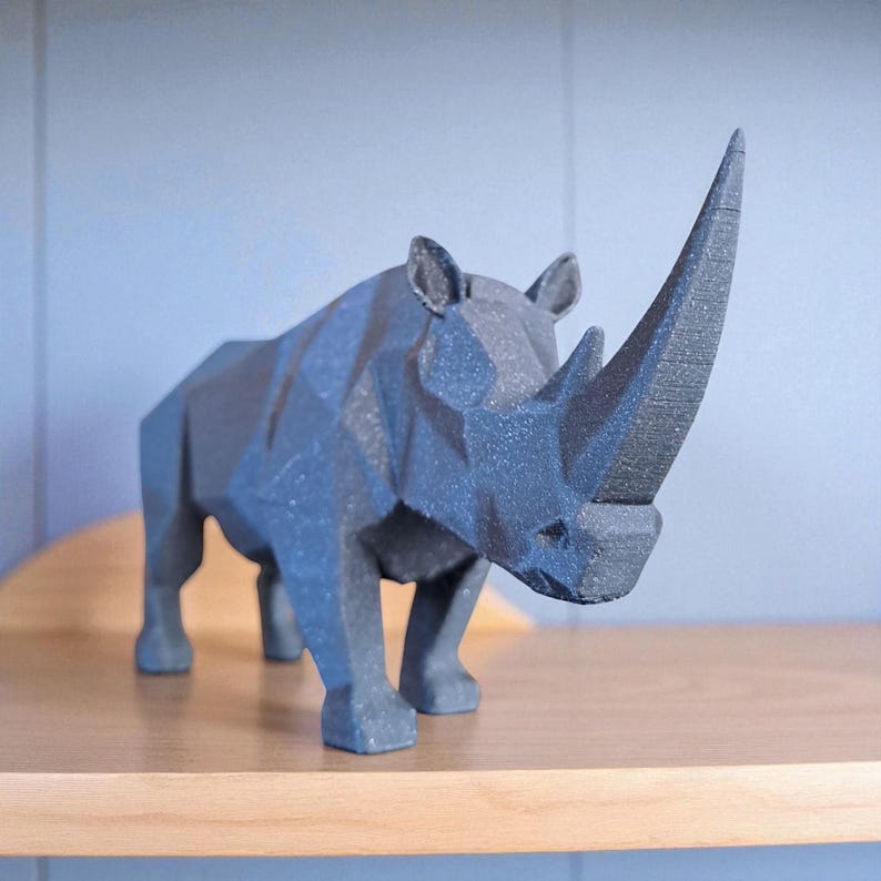 May include: A dark gray, geometric rhino sculpture with a prominent horn. The rhino is positioned on a light brown wooden shelf against a light blue wall. The sculpture's design features sharp angles and a textured surface, giving it a modern aesthetic.