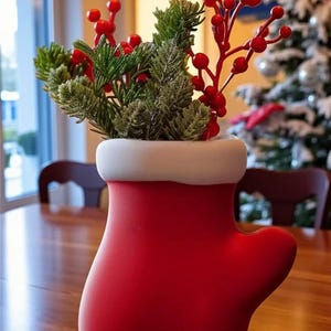 May include: A red mitten-shaped vase with a white rim, filled with green and red decorative sprigs. The vase is sitting on a wooden table. A Christmas tree is visible in the background.