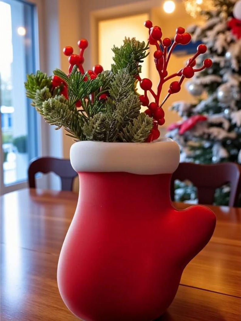 May include: A red mitten-shaped vase with a white rim, filled with green and red decorative sprigs. The vase is sitting on a wooden table. A Christmas tree is visible in the background.