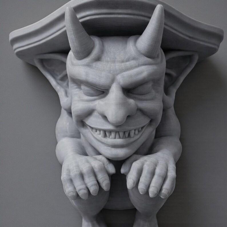 K&ouml;nnte beinhalten: Eine graue, 3D-gedruckte Gargoyle-Regalhalterung mit einem l&auml;chelnden Gesicht, H&ouml;rnern und ausgestreckten H&auml;nden. Das Regal hat eine dekorative Oberkante und ist f&uuml;r die Wandmontage konzipiert. Der Wasserspeier hat detaillierte Gesichtsz&uuml;ge und eine strukturierte Oberfl&auml;che.